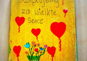 Na zdjęciu deska kuchenna pomalowana na żółto. Na niej napis Dziękujemy za wielkie serce, dookoła czerwone, namalowane serca i wazon z kolorowymi kwiatami. Na dole pieczątka szkoły.