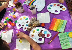 Na zdjęciu stół przykryty fioletową folią. Przy stole siedzą dzieci, malują farbami na kafelkach. Na stole leżą kafelki, kubeczki z woda, palety z farbami i pędzelki. Na kafelkach namalowane są kolorowe obrazki.