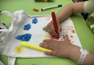 Na zielonym stole leżą ręce dziecka rysującego flamastrami na płóciennej torbie. Rysunek przedstawia dziewczynkę w czerwonej sukience, nad nią świeci żółte słoneczko i pływają niebieskie chmurki.