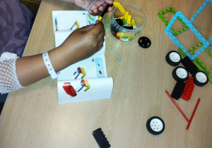 Przy beżowym stole siedzą dwie osoby. Na stole leżą rozrzucone klocki lego. Dziecko buduje coś z klocków lego, według instrukcji.