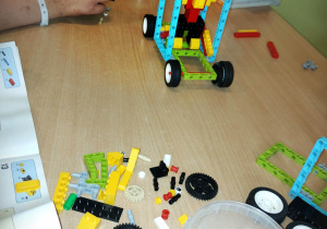 Na beżowym stole leżą klocki lego, instrukcja i gotowa praca z nich zbudowana. Przedstawia ludzika w wozie, podnoszącego sztangę.