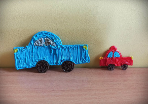 Na zdjęciu dwa samochody wykonane długopisem 3 D. Większy jest niebieski, ma czarne kola i szare szyby. Mniejszy jest czerwony, ma niebieskie szyby i czarne koła.