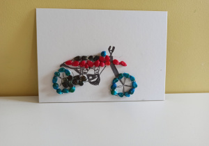 Na jasnej kartce przyklejone kolorowe ziarenka kukurydzy. Z ziarenek ułożony jest obrazek przedstawiający czerwono - niebieski motocykl.