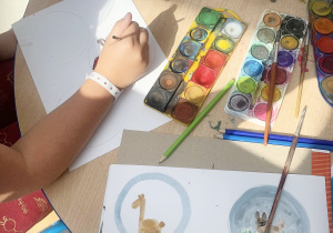 Na okrągłym, beżowym stole leżą farby, pędzle, ołówki i woda do farb w kubeczku. Przy stole siedzą dzieci i malują na kartkach papieru w kółkach, wymyślone przez siebie obrazki .