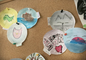 Do korkowej tablicy przyczeppione są kółka wycięte z papieru, a na nich namalowane są przez dzieci wymyślone przez nich rysunki. Wsród nich widzimy: sowę, dinozaura, kąpiącego się pieska, kota, serduszko zachód słońca, jesienne drzewo.