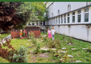 W ogródku przed budynkiem szpitala spacerują dwie dziewczynki wśród zasadzonych roślin.