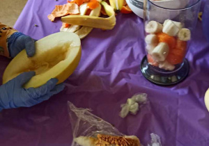 Stół przykryty fioletową folią a na nim biała miska z owocami, obierki owoców na folii, blender z bananami, mandarynkami, na folii pestki dyni. Przy stole stoi dziecko z kawałkiem dynii.