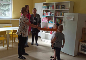 Żóta sala z białymi meblami. W niej stoją trzy uśmiechnięte panie. Jedna z nich pasuje dwoje dzieci na uczniów.