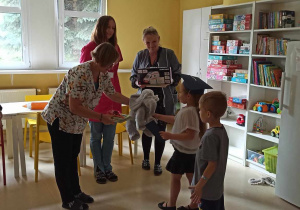 Żółta sala w której stoją białe półki z puzzlami. Stoi w niej 6 pań. Jedna z nich wręcza świeżo pasowanym na uczniów dzieci upominki.