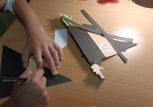Dziecko wykonuje z papieru dwie czarownice w czarnych strojach lecące na miotłach.