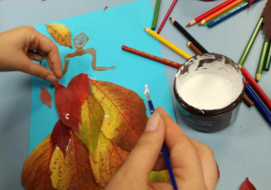 Na szarym blacie widać klej w pojemniczku, rozsypane kolorowe kredki i niebieską kartkę do której dziecko przykleja kolorowe, jesienne liście, tworząc portret Panni Jesieni.