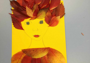 Na szarym blacie leży żółta kartka na której, widać kobiecy portret. Kobieta ma ubranie i włosy wykonane z jesiennych liści.