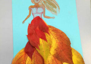 Na szarym blacie leży niebieska kartka na której wykonana jest praca plastyczna. Praca przedstawia kobietę narysowaną kredkami. Suknię ma wyklejoną jesiennymi liśćmi w ognistym kolorze .
