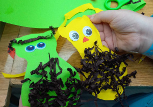 Dziecko trzyma wycięte kształty jeży w żółtym i zielonym kolorze. Jeże mają przyklejone oczy i gły z karbowanych pasków brązowego papieru.