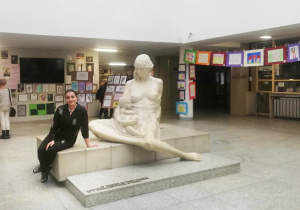 W korytarzu Szpitala Centrum Zdrowia Matki Polki siedzi na podeście z rzeźbą Kobiety z dzieckiem, pani. Wokół rozwieszone są prace dzieci