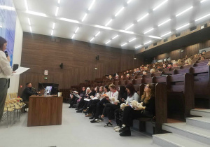 Sala konferencyjna. Z lewej strony siedzi przy biurki prelegent i stoi kobieta mówiąca do mikrofonu. Z prawej strony widać siedzących uczestników konferencji