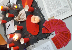 Na białym stole leży czarna chusta z różnokolorowymi pasami. Na niej leżą czerwone , jednorazowe kubki, rozłożone w wachlarzyk karty z czerwonymi koszulkami, dwie świece, lampion i kartki z wróżbami.