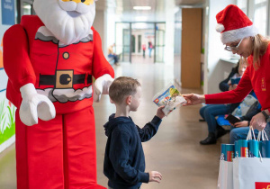 W korytarzu szpitalnym Śnieżynka daje prezent , klocki lego, małemu chłopcu, czemu przygląda się duży Mikołaj LEGO.