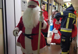 Korytarzem szpitala idzie św. Mikołaj. Za nim stoi kilka osób, wśrod nich strażak w slużbowym stroju.