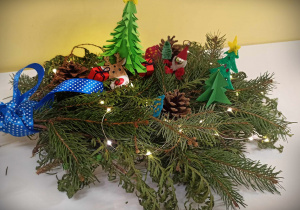 Stroik świąteczny wykonany z gałązek jodły. Stroik jest ozdobiony reniferem, Mikołajem, choinkami zrobionymi z papieru i szyszek
