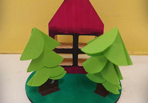 Na krążku zielonego papieru przyklejone są dwie choinki i kartonowy domek z czerwonym dachem.