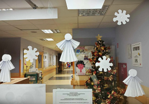 Na przeszklonych drzwiach oddziału wiszą białe śnieżynki i białe aniołki wykonane z papieru. W tle widać szpitalny korytarz.