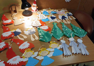 Na blacie stołu leżą ozdoby świąteczne wykonane przez dzieci. Wśród nich są choinki , Mikołaje, aniołki.