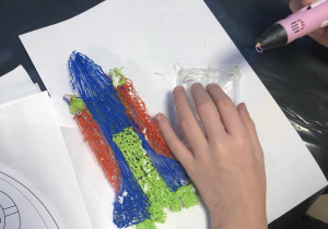 Na zdjęciu widać rękę dziecka wykonującą na białej podkładce czerwono-niebiesko-zieloną rakietę kosmiczną długopisem 3D.