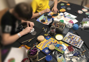 Dzieci siedzą przy stole przykrytym czarną folią i wykonują prace plastyczne. Na stole leżą materiały plastyczne, takie jak farby, nożyczki, kredki, naklejki.