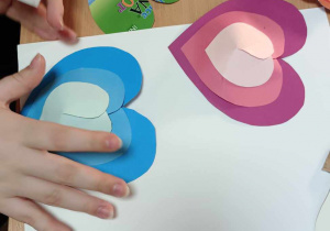 Na stole leży biała kartka do krórej dziecko przykleja dwa trójkolorowe sedduszka 3D.