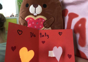 Dziewczynka trzyma zabawkowego bobra z czerwonym serduszkiem , i czerwoną laurkę z przyklejonymim żółtym i różowym serduszkiem. Markerem narysowane na niej są serduszka i napis tata.