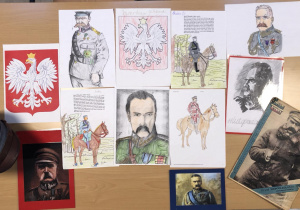 Na beżowym blacie stołu leżą karty pracy rysunek i gazetki ze zdjęciami Józefa Piłsudskiego.