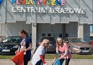 Trzy panie stoją przed budynkiem Centrum Urazowego Instytutu Centrum Zdrowia Matki Polki i zbierają z trawnika śmieci do dużych worków foliowych.