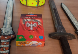 Korona koloru żółtego wykonana z papieru ozdobiona kolorowymi kryształkami. Na środku zdjęcia jest widoczna gra zatytułowana "Historia Polski" z wizerunkiem godła Polski oraz leżące po obu stronach atrapy mieczy