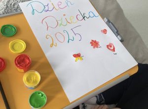 Na szpitalnym łóżku, na blacie, znajduje się biała kartka z kolorowym napisem „Dzień Dziecka 2025” oraz rysunkami: serca, kwiatka i motyla. Obok kartki widoczne są farby w kolorach: żółtym,