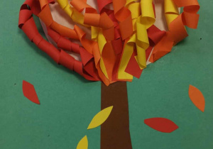 Na ciemnozielonym kartonie z kolorowego papieru naklejono drzewo - brązowy pień i koronę, którą stanowią pomarańczowe i żółte liście powstałe ze skręcenia papieru. Nad drzewem białe i niebieskie chmurki, a pod nim zielona trawa mocno ponacinana. Praca jest przestrzenna.