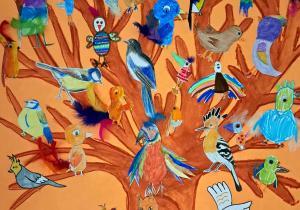 Na dużym pomarańczowym kartonie narysowane jest brązowe drzewo z licznymi gałęziami. Doklejone są na nim różne ptaki wykonywane mieszanymi technikami plastycznymi.