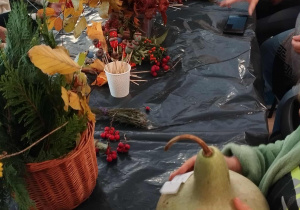 Długi stół zabezpieczony czarną folią. W koszyczkach ustawione suszone rośliny, owoce, ozdobne dynie.