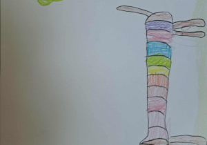 Na zdjęciu znajduje się rysunek wykonany kredkami na białej kartce. Przedstawia psa o bardzo długim, smukłym ciele, które jest podzielone na kolorowe pasy (różowy, fioletowy, pomarańczowy, zielony, niebieski). Pies ma prostą sylwetkę, czarny nos i zaznaczone oko. Obok, po prawej stronie, narysowane jest drzewo z brązowym pniem i zieloną koroną. Tło jest puste, co sprawia, że głównymi elementami rysunku są pies i drzewo. Całość wygląda na dziecięcą, kreatywną pracę plastyczną.