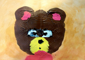 Obraz przedstawia kompozycję wykonaną z masy plastycznej (prawdopodobnie plasteliny) na tle w odcieniach jasnego pomarańczu i beżu. Centralnym elementem jest duża, stylizowana głowa misia.