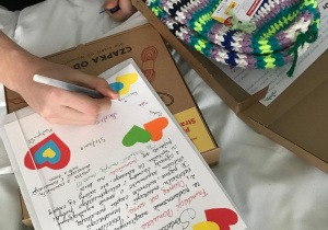 Na zdjęciu widać rękę osoby piszącej długopisem na kartce ozdobionej kolorowymi serduszkami. Kartka zawiera odręczne notatki lub dedykacje. Obok znajduje się otwarte pudełko, w którym leży kolorowa, ręcznie robiona czapka w paski (zielone, fioletowe, białe, czarne i żółte kolory) z metką i medalikiem. Całość wygląda na element akcji charytatywnej lub prezentu dla kogoś, np. pacjenta w szpitalu.