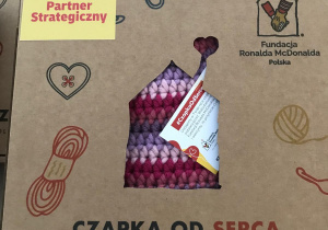 Na zdjęciu widać pudełka z napisem „Czapka od serca – dla Ciebie od wolontariuszy”, przygotowane przez Fundację Ronalda McDonalda Polska we współpracy z DHL Express (Partner Strategiczny). W opakowaniu znajduje się ręcznie robiona, kolorowa czapka na drutach, widoczna przez wycięty otwór w kartonie. Grafika na pudełku przedstawia motywy związane z szyciem i robótkami ręcznymi (nici, nożyczki, serduszko).