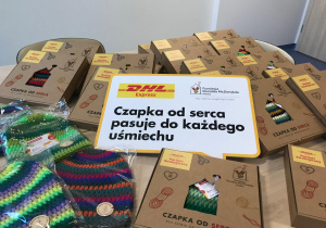 Na zdjęciu widać stół pełen pudełek z napisem „Czapka od serca – dla Ciebie od wolontariuszy”, przygotowanych przez Fundację Ronalda McDonalda we współpracy z DHL Express. Na pierwszym planie znajdują się kolorowe, ręcznie robione czapki w paski (zielone, fioletowe, czerwone, żółte), a obok duża tabliczka z hasłem: „Czapka od serca pasuje do każdego uśmiechu”.
