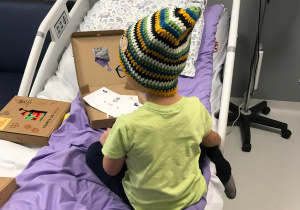 Na zdjęciu widać dziecko siedzące na łóżku szpitalnym, ubrane w kolorową, ręcznie robioną czapkę w paski (zielone, żółte, białe, granatowe). Obok znajdują się pudełka z napisem „Czapka od serca” oraz kartka z ozdobami, prawdopodobnie element akcji charytatywnej.