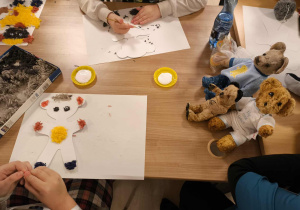 Na zdjęciu widać stół, na którym odbywają się zajęcia plastyczne. Na kartkach leżą wycięte sylwetki misiów, które są ozdabiane kolorowymi elementami z futerka i bibuły. Obok znajdują się małe pojemniczki z klejem, pudełko z materiałami dekoracyjnymi oraz kilka pluszowych misiów w ubrankach.