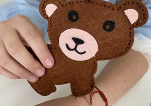 Na zdjęciu widać rękę dziecka trzymającą małego, ręcznie wykonanego misia z filcu. Miś jest brązowy, ma różowe elementy na pyszczku i uszach, czarne oczy oraz uśmiechnięty pyszczek z noskiem. Brzegi misia są starannie przeszyte ozdobnym ściegiem.