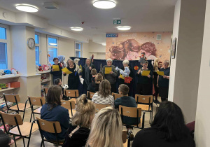 Na zdjęciu widać salę, w której odbywa się występ artystyczny. W tle stoi grupa osób trzymających kolorowe kartki, prawdopodobnie śpiewających lub recytujących tekst. Niektóre osoby trzymają pacynki lub maskotki, co sugeruje, że jest to przedstawienie dla dzieci. Na pierwszym planie siedzi publiczność – kilka rzędów krzeseł zajętych przez dzieci i dorosłych. W pomieszczeniu są jasne ściany, duże okna, a na ścianie w tle widoczny jest baner z logo „pepco” i grafiką przedstawiającą postacie.