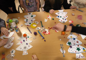 Na zdjęciu widać stół, na którym odbywają się kreatywne zajęcia plastyczne. Na blacie rozłożone są papierowe choinki wycięte z białego kartonu, które dzieci ozdabiają kolorowymi elementami – naklejkami w kształcie gwiazdek, pomponikami, cekinami, koralikami i brokatem. Widać też klej w małych żółtych pojemniczkach, kolorowe pisaki, arkusze z samoprzylepnymi ozdobami oraz butelkę wody. Przy stole siedzi kilka osób, które pracują nad dekoracjami, a w tle widać fragment sali.