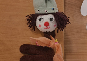 Na zdjęciu znajduje się ręcznie wykonana pacynka w formie postaci klauna lub lalki. Głowa jest zrobiona z białej kulki styropianowej, z namalowanymi oczami, czerwonym noskiem i różowymi policzkami. Włosy wykonane są z brązowej włóczki, a na głowie znajduje się jasnozielony materiałowy kapelusz ozdobiony kolorowymi koralikami i przewiązany zieloną włóczką. Tułów pacynki to brązowa rękawiczka, a wokół szyi owinięta jest pomarańczowa apaszka z małymi koralikami.