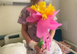 Dziecko stoi w szpitalnym pokoju i trzyma w rękach dużego, kolorowego kwiatka zrobionego z bibuły. Kwiat ma różowe i żółte płatki oraz zieloną łodygę. Dziewczynka ma na sobie fioletową koszulkę z jednorożcem i tęczą, a na lewej dłoni widoczny jest opatrunek, co sugeruje niedawny zabieg lub pobranie krwi. W tle widać szpitalne łóżko z poduszką oznaczoną napisem „UM w Łodzi”.